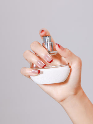 Hand holding a perfume bottle wearing nude press-on nails with metallic red French tips