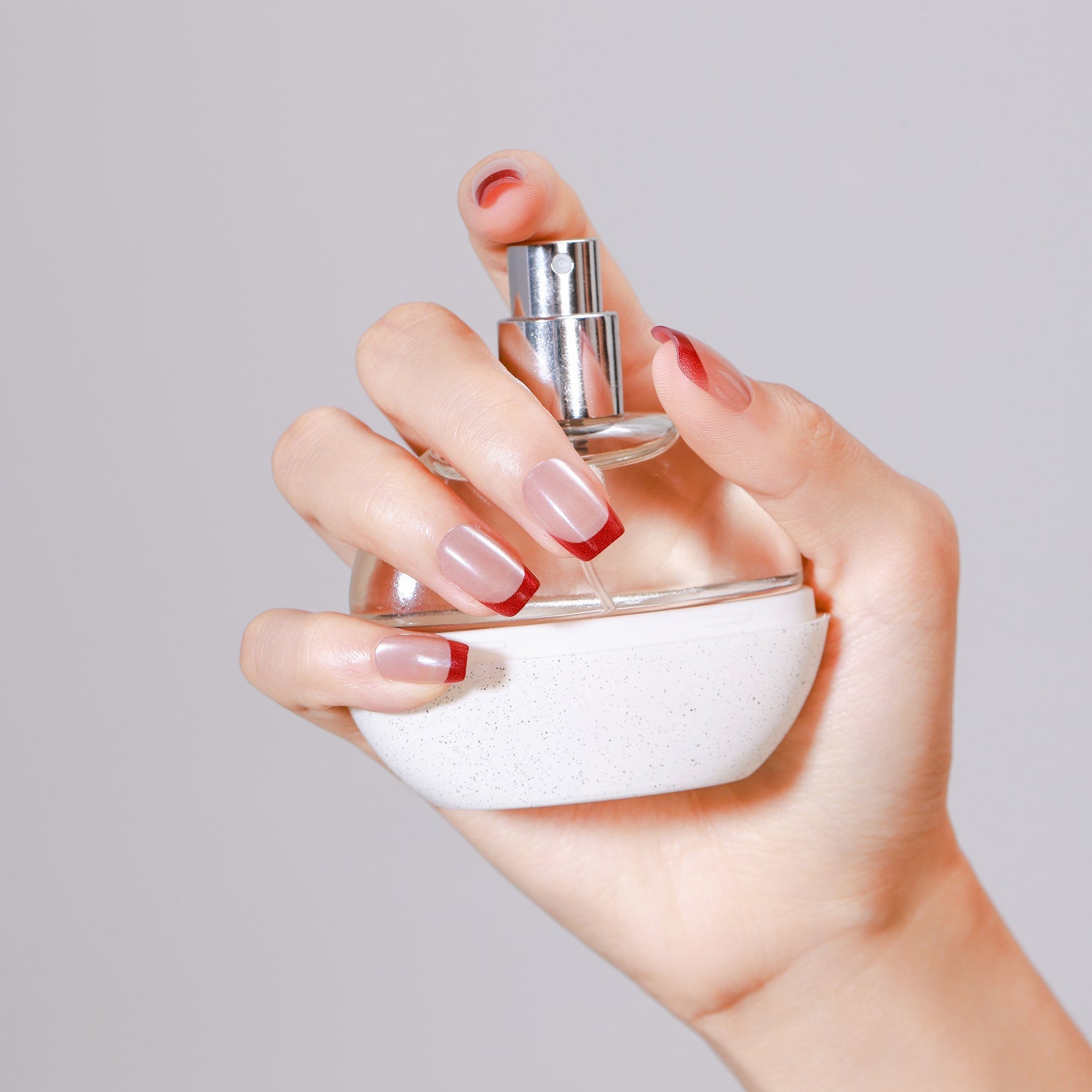 Hand holding a perfume bottle wearing nude press-on nails with metallic red French tips