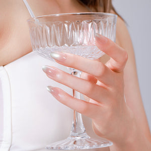 Hand holding glass wearing Satin Sands long oval champagne nude shimmer press-on nails