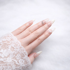 Almond press-on nails with nude base and white French tips, rhinestone accent on ring finger and white swirl design, photographed on a model hand with white lace sleeve in studio lighting