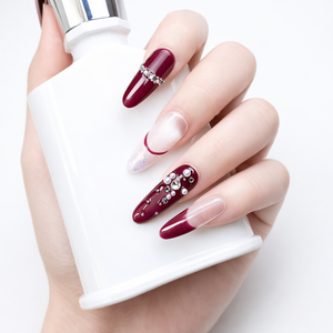 Hand model holding a white bottle while wearing burgundy French press on nails with nude ombre base and crystal rhinestone accents