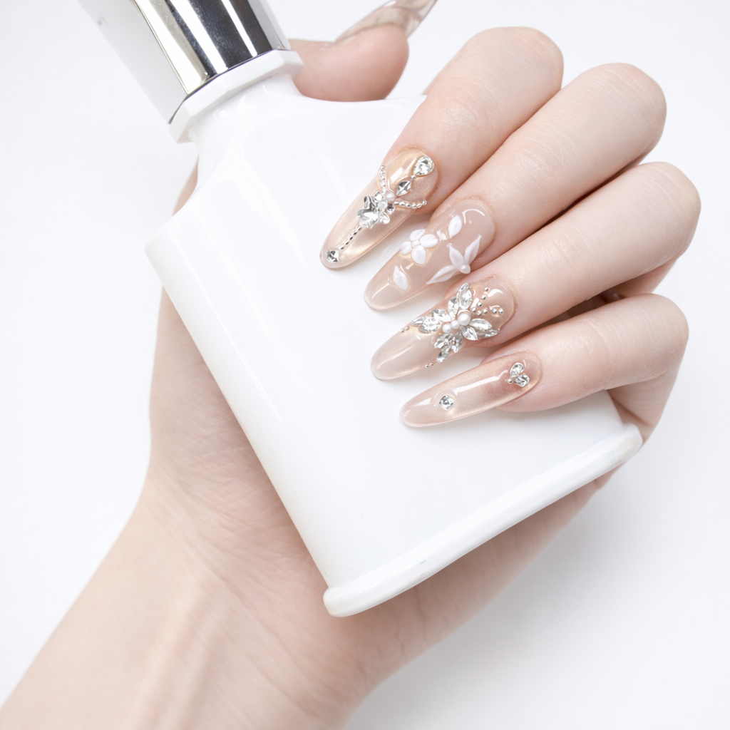 Champagne nude almond press-on nails with crystal flower and rhinestone accents, shown on a hand holding a white cosmetic bottle.
