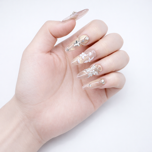 Hand model wearing champagne nude almond press on nails with silver crystal charms, floral details and a soft glossy neutral finish