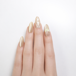 Professional studio photo of a white skin model hand wearing gold champagne almond press-on nails, featuring a quilted pearl accent nail and a nude sheer nail with a pale yellow bow design on a clean white background.