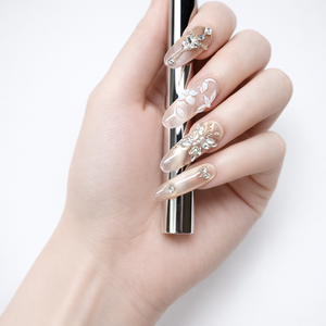 Nude almond press-on nails featuring crystal cross and floral embellishments, photographed on a hand holding a chrome cylinder against a white background.