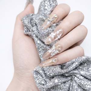 Close-up of nude champagne almond press-on nails with crystal floral accents and silver rhinestone details, styled on a hand holding glitter fabric.