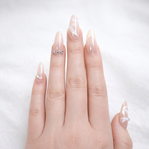 Professional white skin model hand wearing almond-shaped French ombre press-on nails with nude pink base, soft white tips, white swirl nail art accents, and a delicate rhinestone ring-finger detail on a clean white studio background.