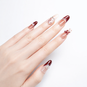 White-skin model hand wearing ShadePax almond press-on nails with glossy burgundy French tips, nude base, red polka-dot accent nail, and 3D bow charm in studio lighting.