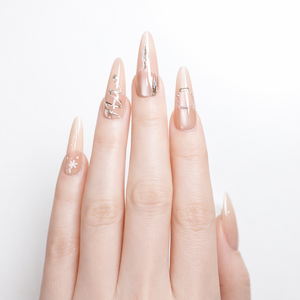 Close-up of nude ombré almond press-on nails with silver chrome swirl accent, vertical silver foil stripe, rhinestone frame detail, and white snowflake nail art on a fair-skin model hand.