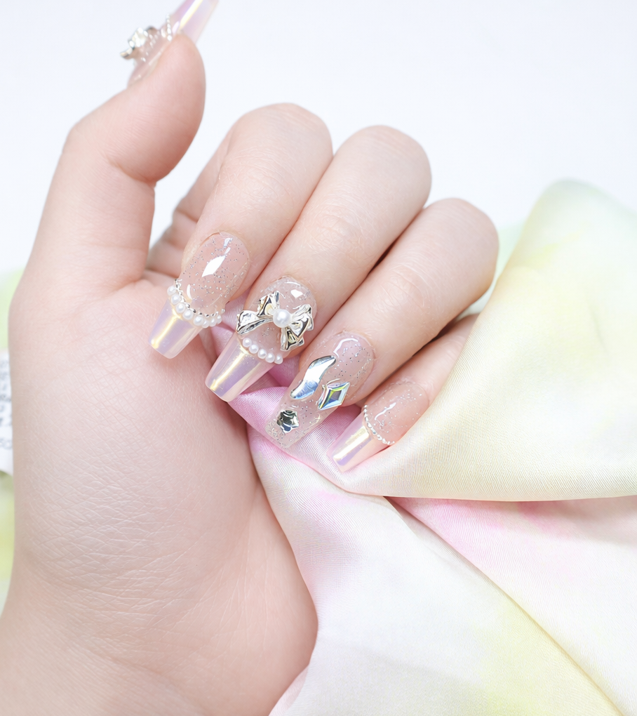 Long coffin press on nails featuring clear glitter overlay, pearlescent chrome tips, pearl beads, metallic bow charms, and crystal accents in close up