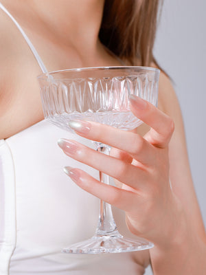 Hand holding glass wearing Satin Sands long oval champagne nude shimmer press-on nails