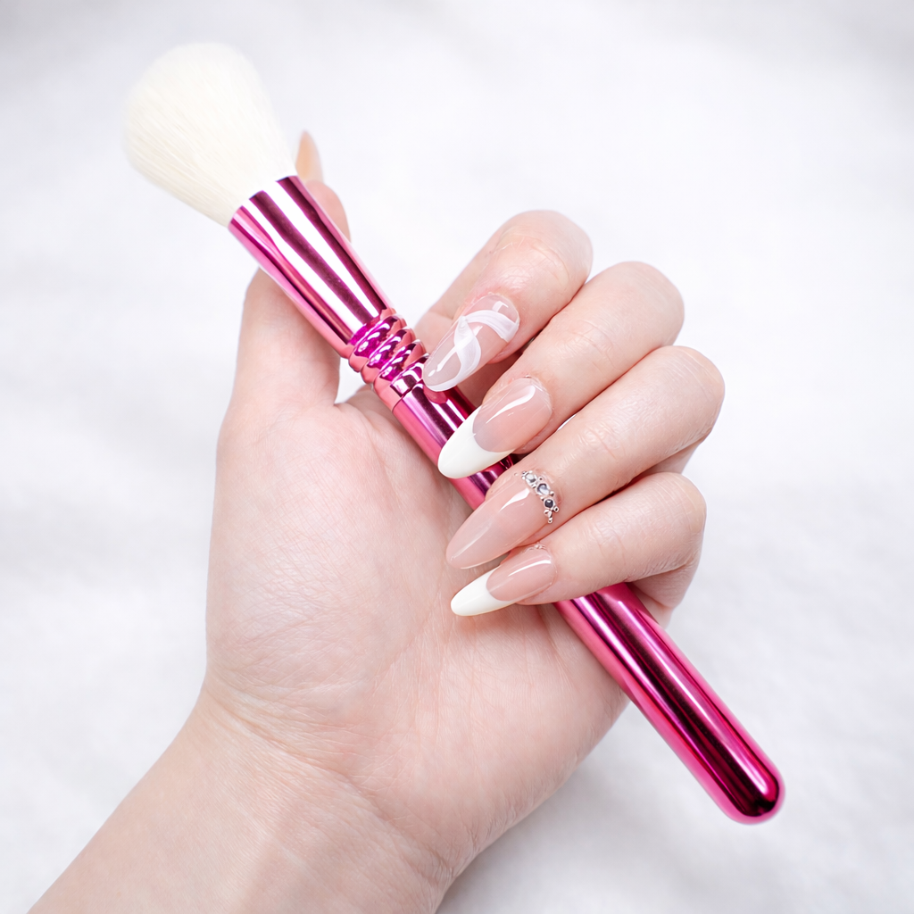 Almond press-on nails with nude base and white French tips, swirl accent nail and rhinestone cuticle detail, holding a pink metallic makeup brush on white studio background