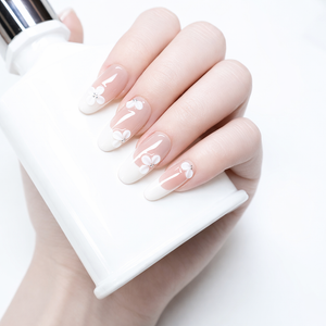 Hand model holding white bottle wearing nude almond French press on nails with white floral accents in a soft bridal style
