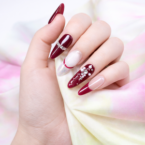 Burgundy French press on nails with nude ombre base and crystal rhinestones shown on hand against a soft pastel fabric background
