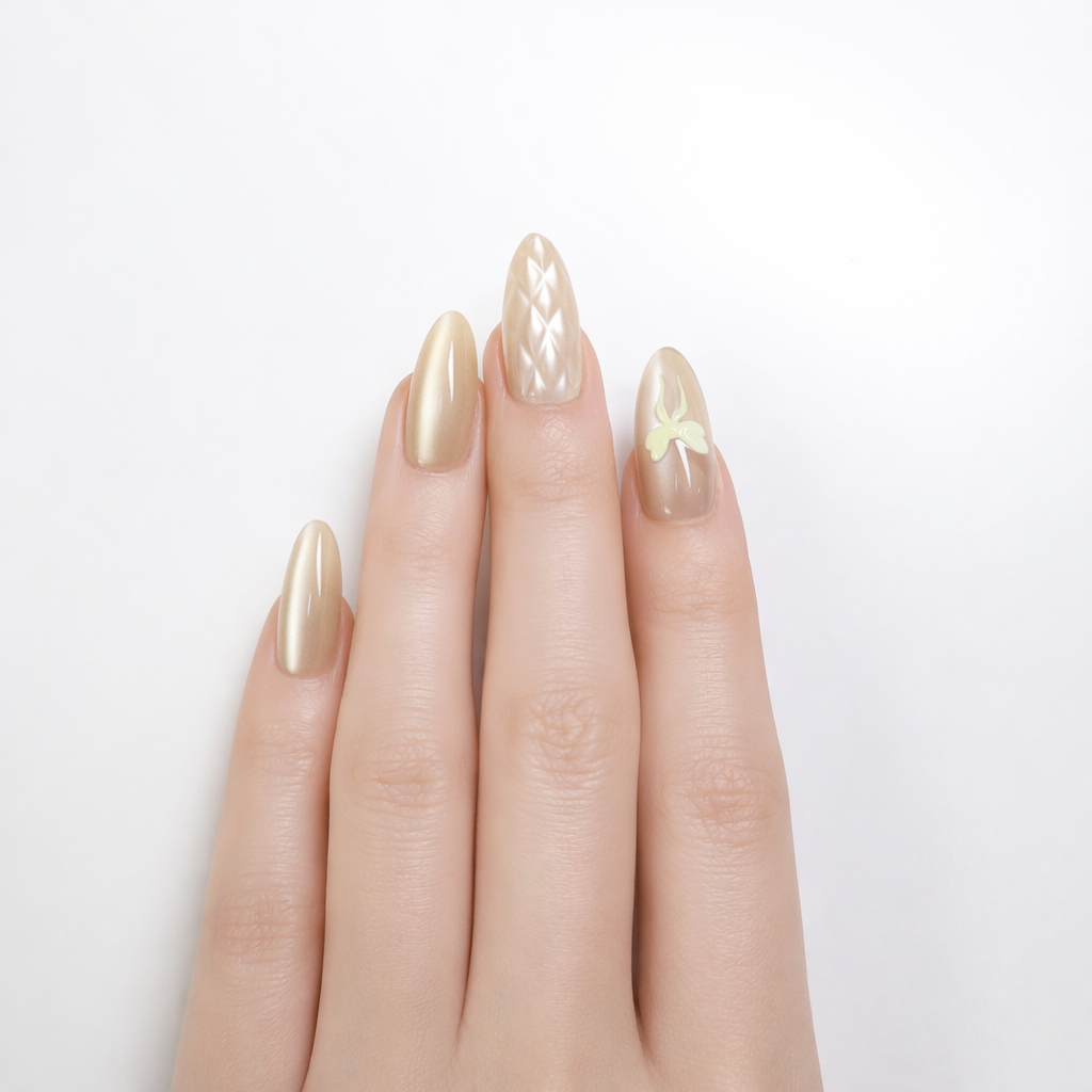 Professional studio photo of a white skin model hand wearing gold champagne almond press-on nails, featuring a quilted pearl accent nail and a nude sheer nail with a pale yellow bow design on a clean white background.