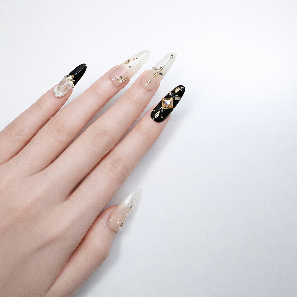 Long almond press-on nails in nude and black with champagne square crystal accent nail, gold foil flakes, rhinestone details, and glossy finish shown on model hand against white background.