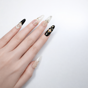 Long almond press-on nails in nude and black with champagne square crystal accent nail, gold foil flakes, rhinestone details, and glossy finish shown on model hand against white background.