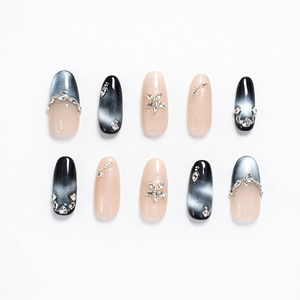 Almond-shaped press-on nails featuring black-to-silver cat-eye ombré tips, glossy nude shimmer accent nails, and clear rhinestone details including a crystal flower centerpiece and gem clusters on a clean white background.