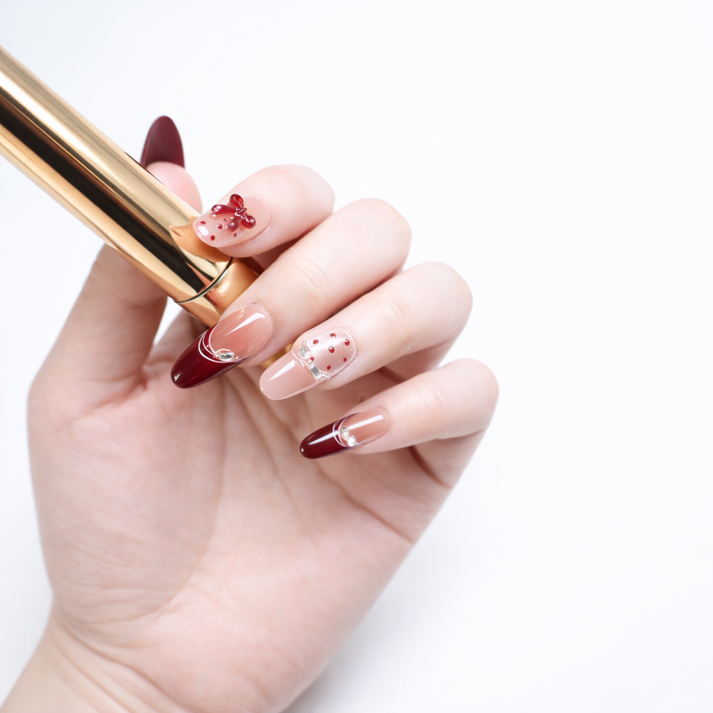 Professional white-skin model hand holding a gold makeup tube, featuring ShadePax almond press-on nails with burgundy French tips, nude base, red polka-dot accent nail, and 3D bow design in studio lighting.