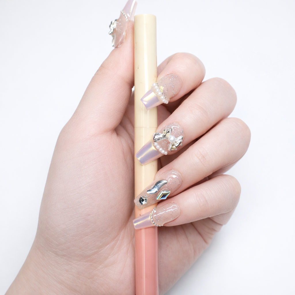 Coffin press on nails with sheer glitter base, pearl edge detailing, silver bow charms, and iridescent crystal embellishments shown holding object