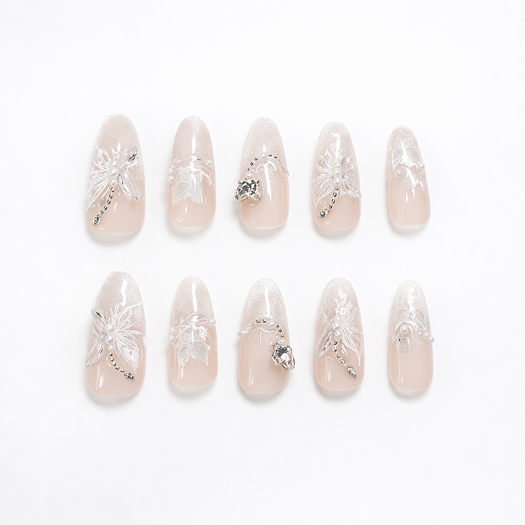Almond-shaped nude-to-ivory ombré press-on nails with a soft shimmer gradient, raised 3D white floral designs, pearl accents, and silver rhinestone/crystal details on a clean white background.