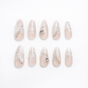 Almond-shaped nude-to-ivory ombré press-on nails with a soft shimmer gradient, raised 3D white floral designs, pearl accents, and silver rhinestone/crystal details on a clean white background.