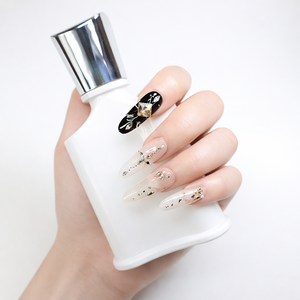 Luxury long almond press-on nails featuring black accent nail with champagne crystal centerpiece, nude ombré base, gold foil, and rhinestone embellishments shown in elegant lifestyle hand pose.