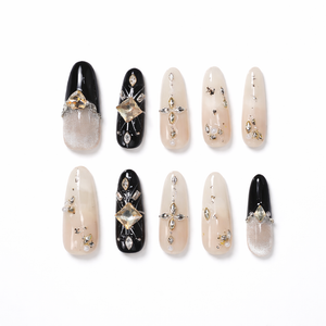 Luxury long almond press-on nails set in glossy black and sheer nude ombré with champagne square crystal accents, gold foil flakes, rhinestone cross details, and pearl embellishments on a clean white background.