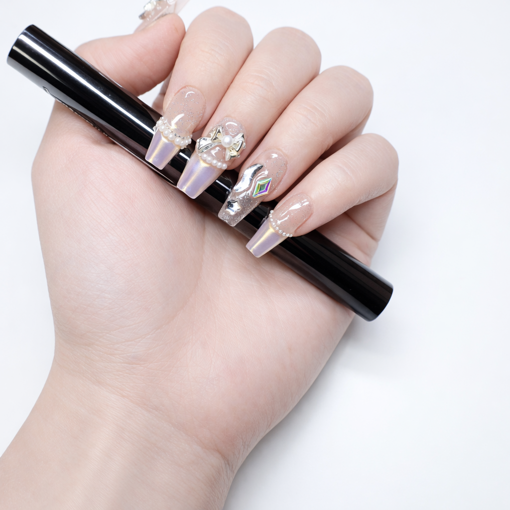 Pearlescent coffin press on nails with clear glitter base, chrome tips, pearl trim, metallic bow nail art, and rainbow crystal accent close up