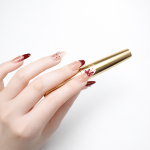 Close-up studio photo of a fair-skin model hand wearing ShadePax almond press-on nails with nude base, deep red French tips, red bow accent nail, rhinestone details, and red dot design, holding a gold tube on a white background.